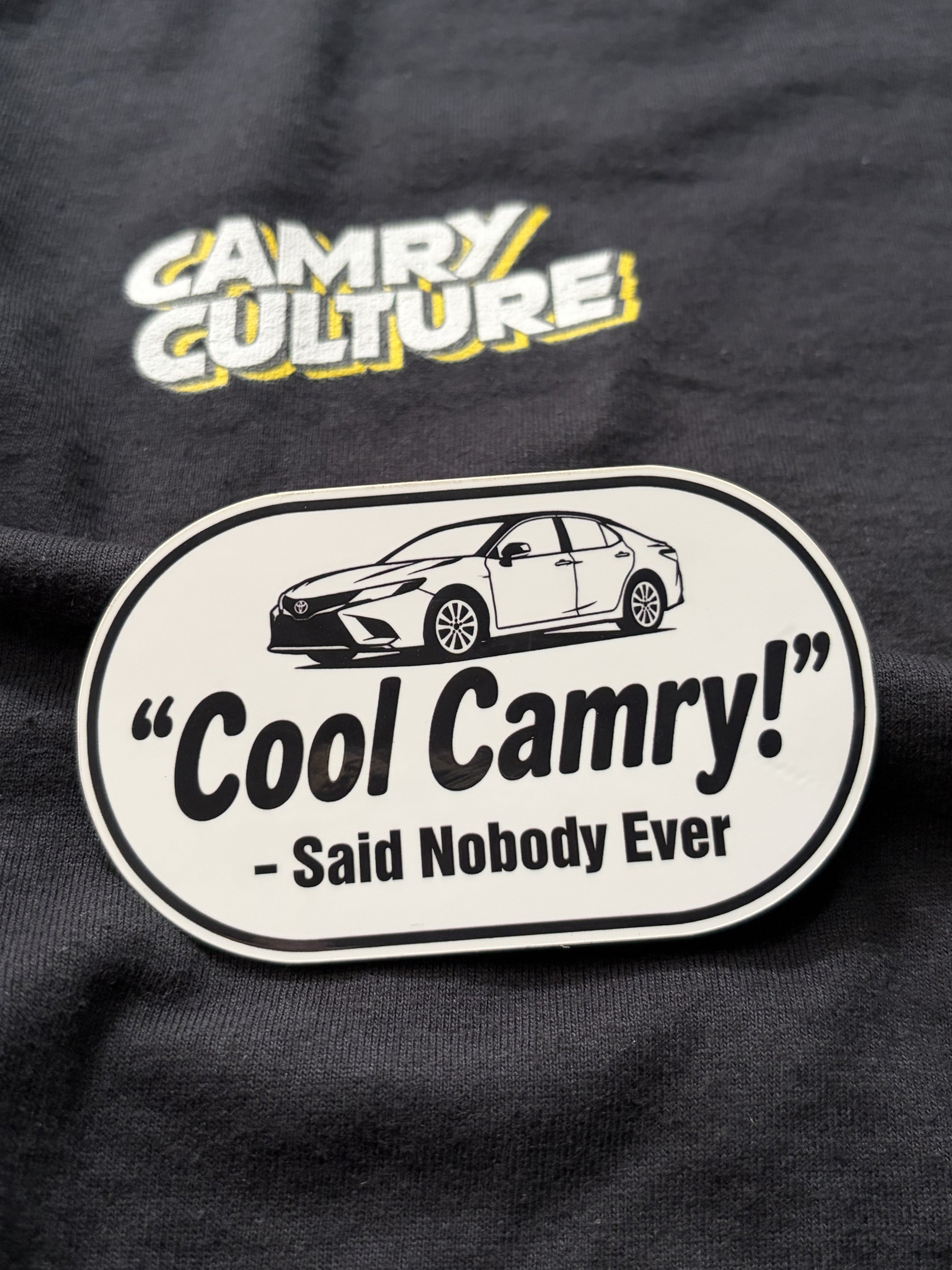 A close-up of a sticker featuring a white and black sticker that reads 'Cool Camry! - Said Nobody Ever' with a car illustration, and a yellow and white 'Camry Culture' graphic above it.