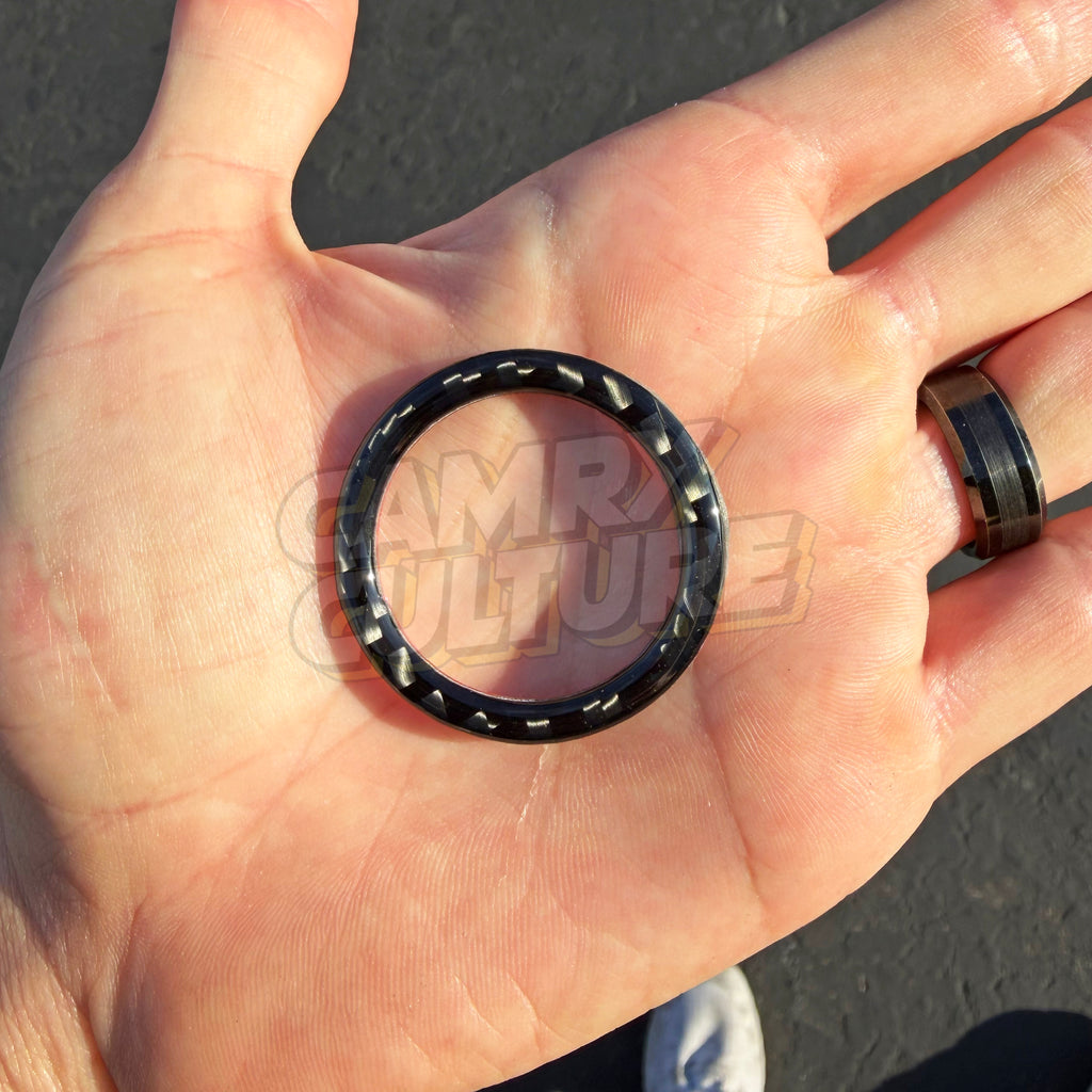 A black carbon fiber ring for a Toyota Camry 2018-2024 with a textured, woven pattern, held in a person's open palm. The person is wearing a matching dark ring on their finger, and the background shows asphalt pavement.