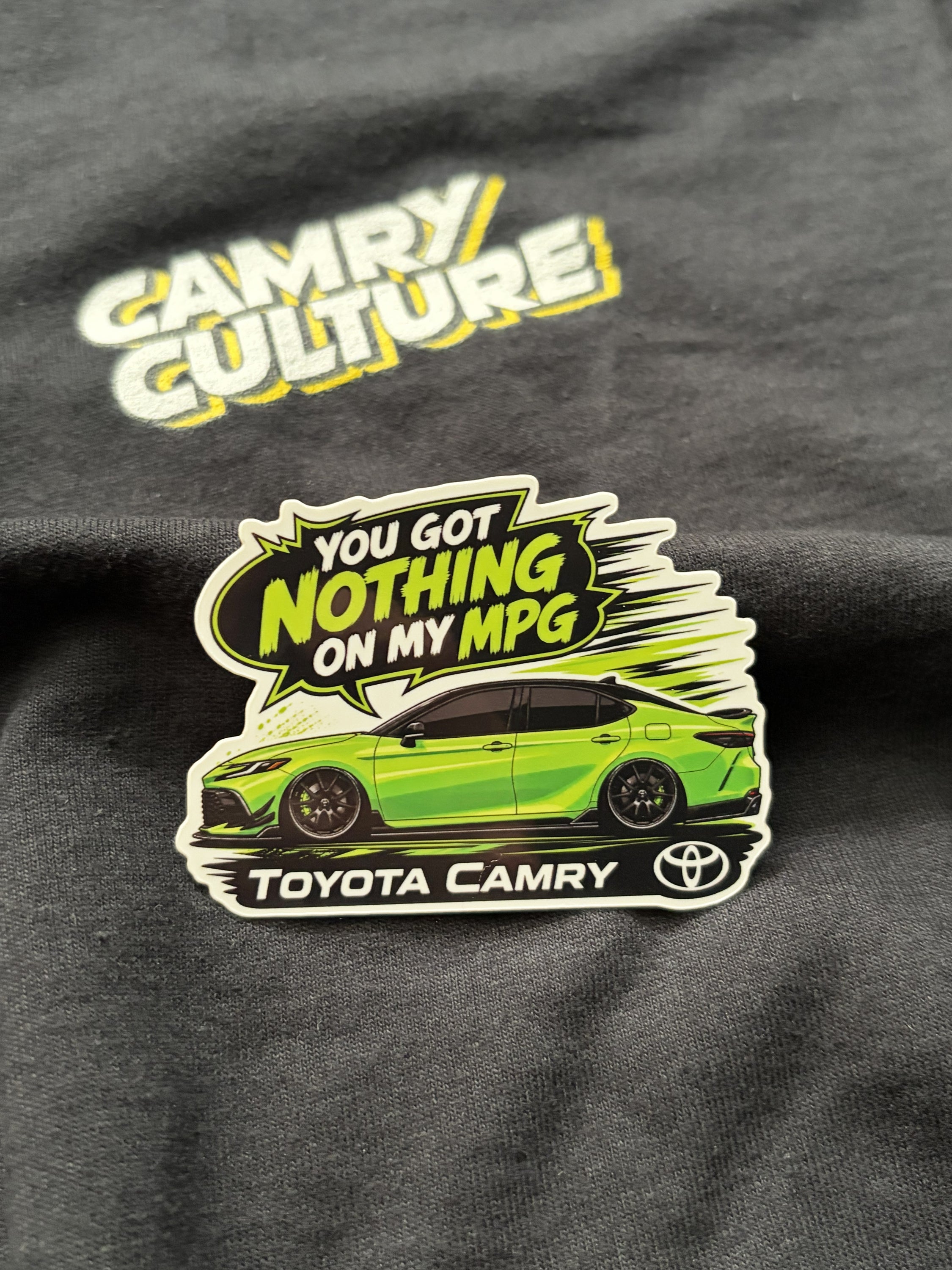 A die-cut sticker featuring a bright green Toyota Camry car and the text 'YOU GOT NOTHING ON MY MPG' with 'TOYOTA CAMRY' and the Toyota logo, displayed on a black t-shirt with a 'Camry Culture' graphic.