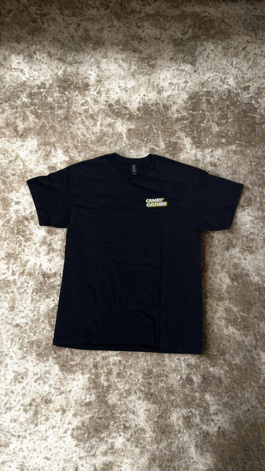 A black short-sleeve t-shirt with a small yellow and white 'CAMRY CULTURE' logo on the left chest, laid flat on a textured gray and beige carpet.