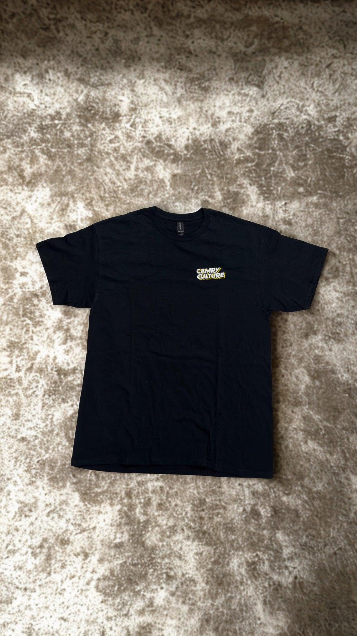 A black short-sleeve t-shirt with a small yellow and white 'CAMRY CULTURE' logo on the left chest, laid flat on a textured gray and beige carpet.