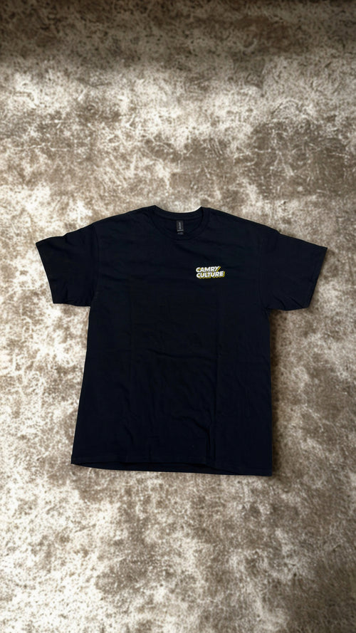 A black short-sleeve t-shirt with a small yellow and white 'CAMRY CULTURE' logo on the left chest, laid flat on a textured gray and beige carpet.