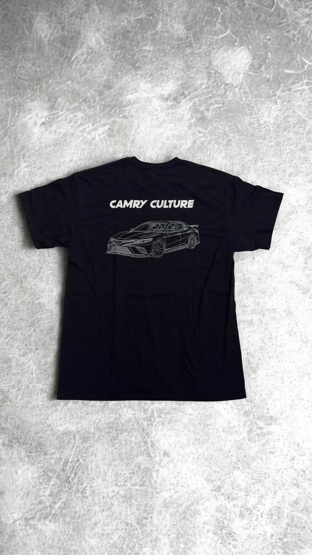 A black short-sleeve t-shirt laid flat on a textured gray concrete background. The back of the shirt features a white graphic print of a sports car and the text 'CAMRY CULTURE'.