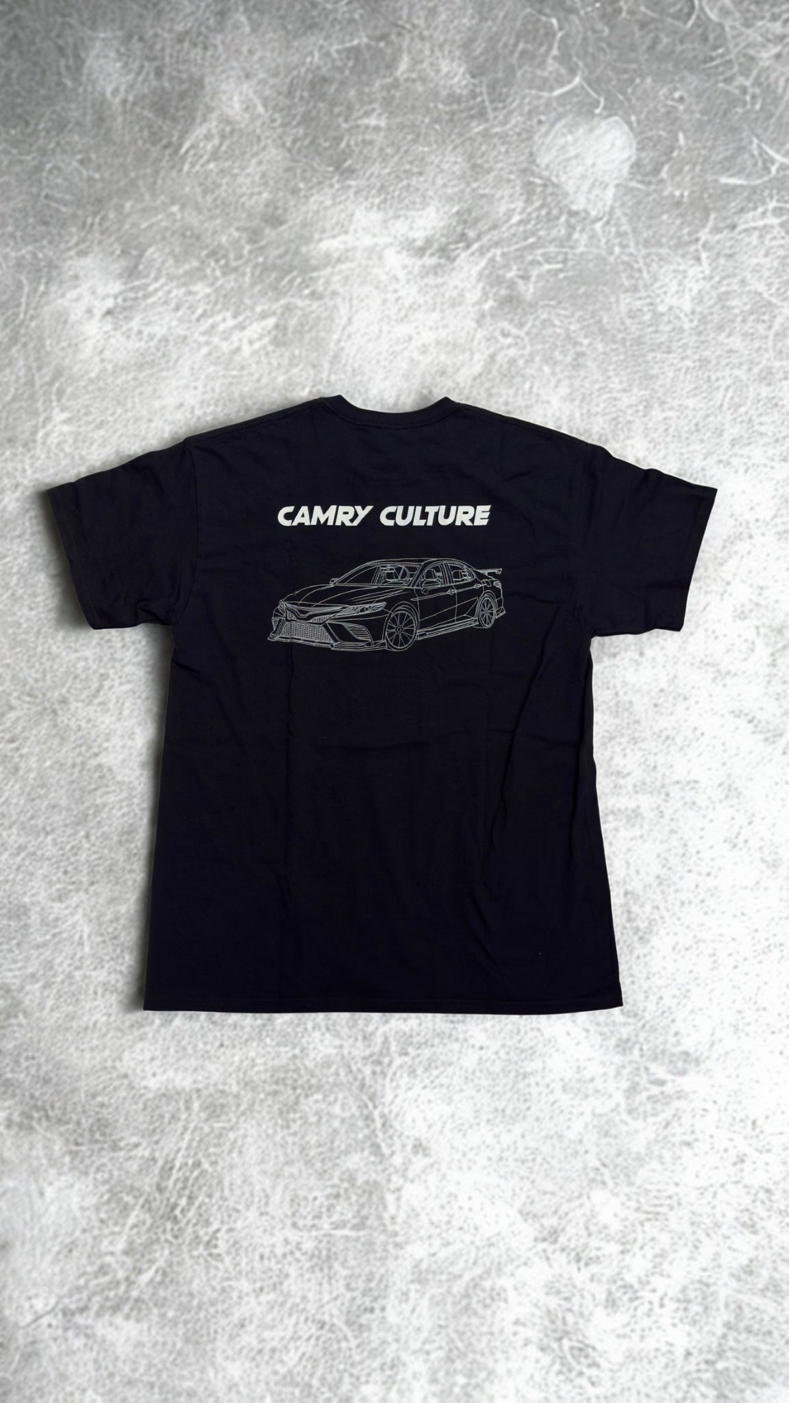 A black short-sleeve t-shirt laid flat on a textured gray concrete background. The back of the shirt features a white graphic print of a sports car and the text 'CAMRY CULTURE'.