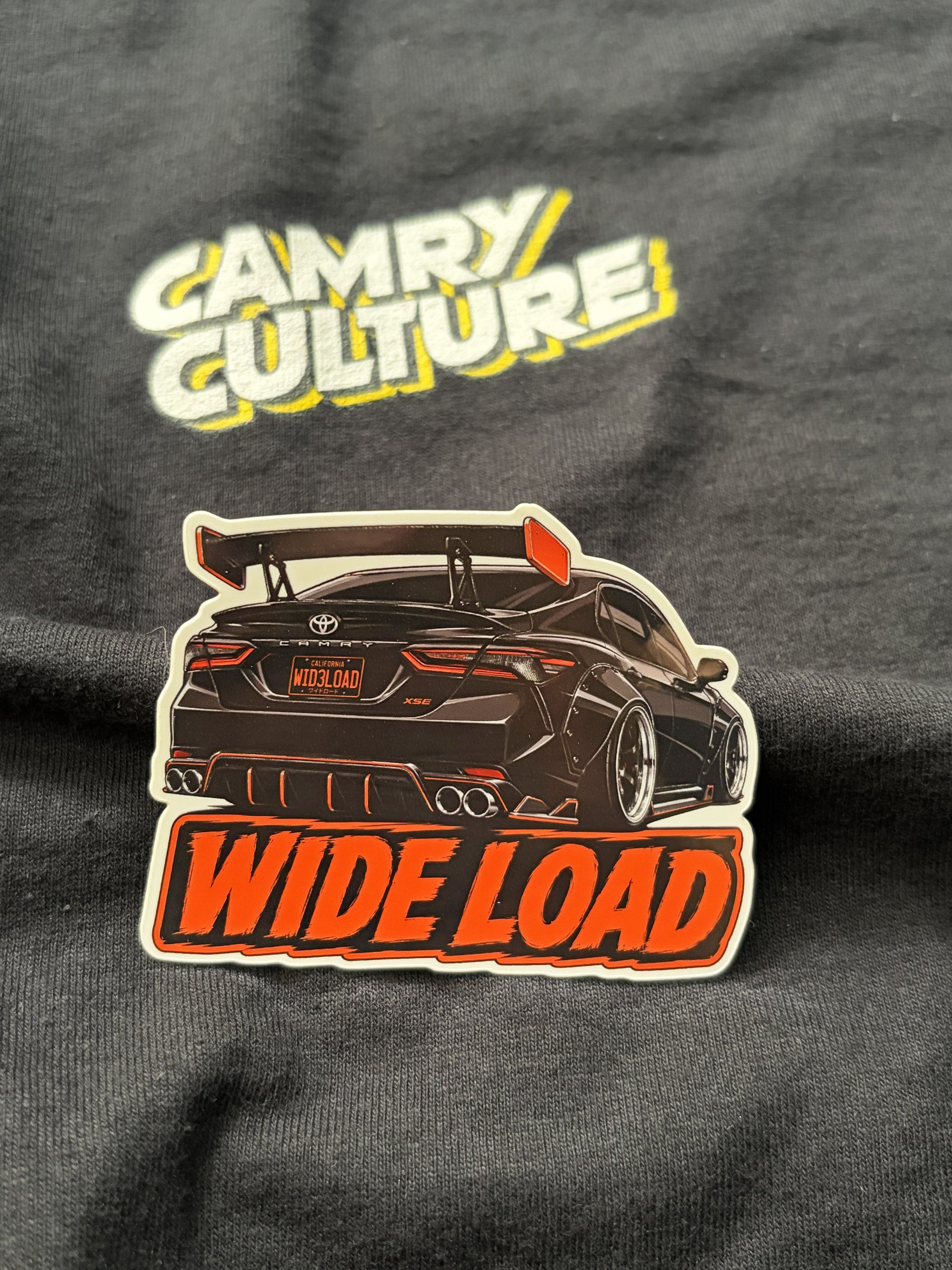 A die-cut sticker featuring a black widebody sports car with orange 'WIDE LOAD' lettering, placed on a dark gray t-shirt with a yellow and white 'CAMRY CULTURE' graphic above it.