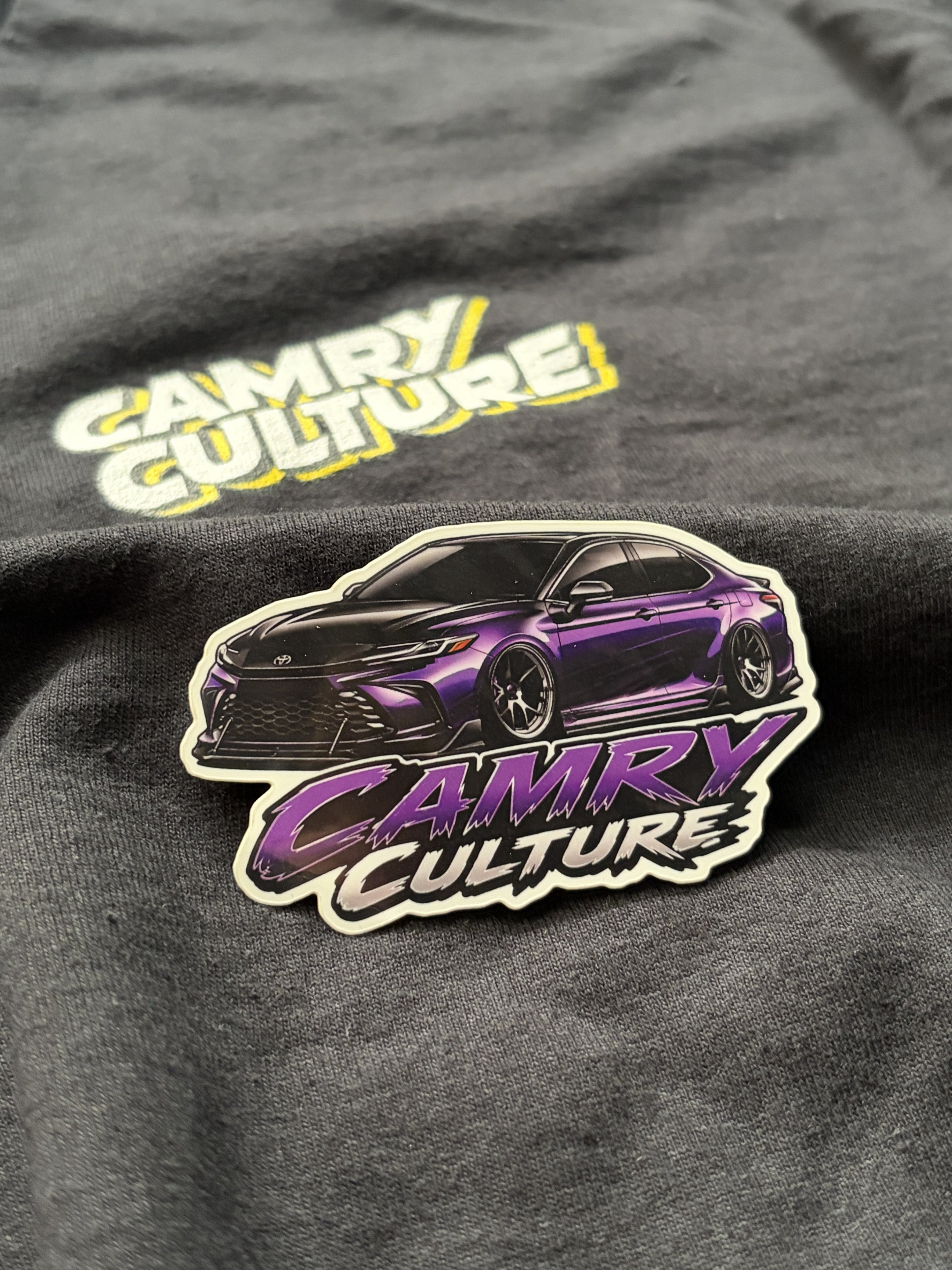 Dark Purple Toyota Camry Sticker that says Camry Culture. Sticker is sitting on top of an official Camry Culture shirt. 