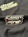 Close up of a "Camry Culture" white retro sticker sitting on top of a black shirt that has the Camry Culture logo. The camry is a XV80 lowered. 