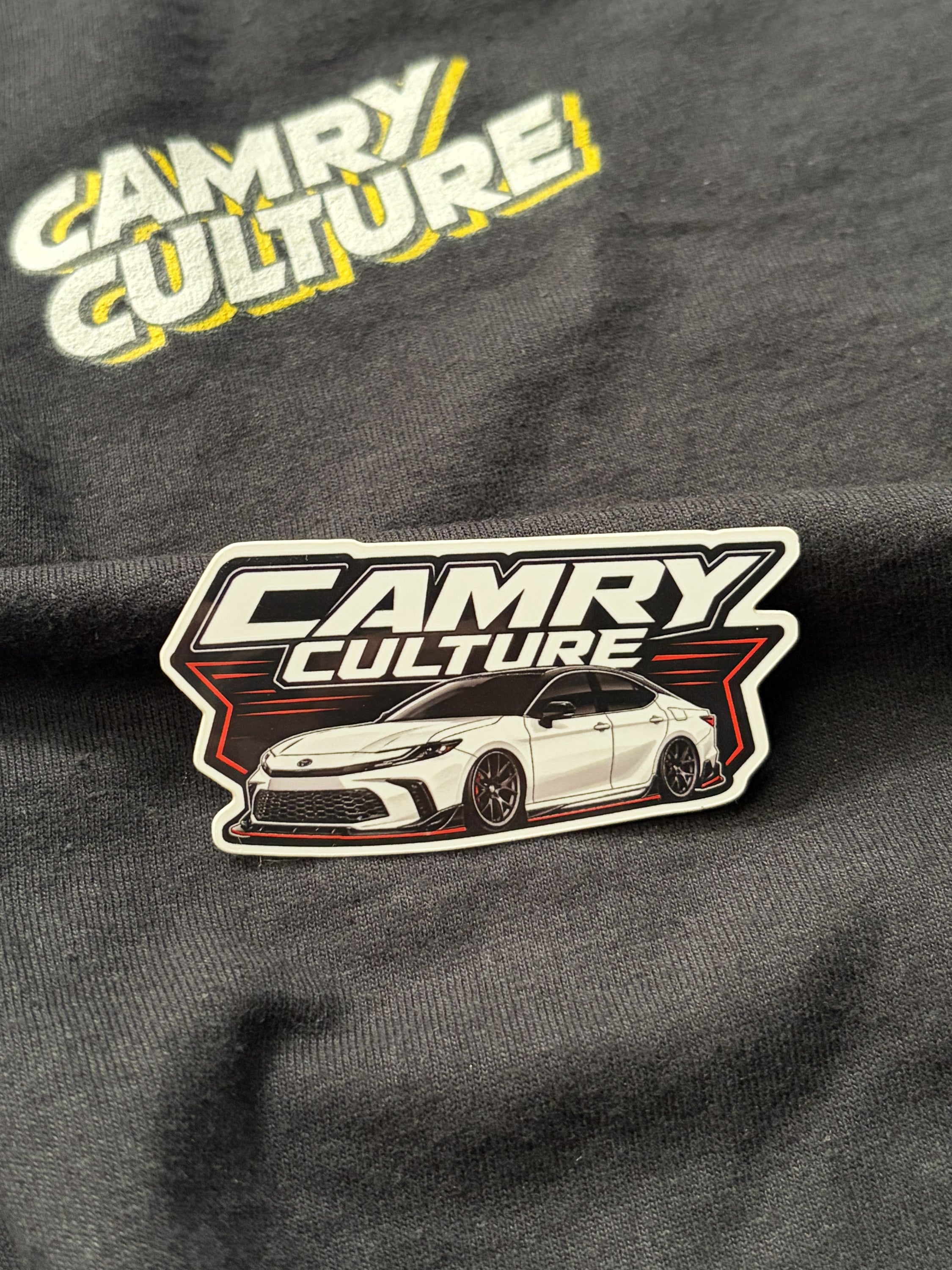 Close up of a "Camry Culture" white retro sticker sitting on top of a black shirt that has the Camry Culture logo. The camry is a XV80 lowered. 